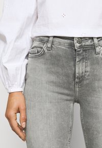 Jeans de brim cinzas com um corte justo, apresentando um design clássico de cinco bolsos e fecho em botão de metal, combinados com uma blusa branca texturizada.