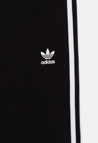 Melni adidas sporta bikses ar baltām sānu svītrām un mazu baltu logotipu, kurā redzama ikoniskā trifeles apzīmējums. Mīksta auduma tekstūra.
