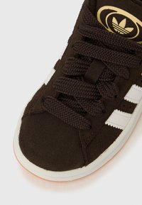 Καφέ σουέντ sneaker με λευκές ρίγες, καφέ κορδόνια με υφή, στρογγυλεμένη μύτη και λαστιχένια σόλα. Λογότυπο στη γλώσσα σε χρυσό χρώμα.