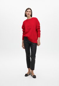Roter Pullover mit geprägtem Logo, rundem Ausschnitt und überschnittenen Schultern. Kombiniert mit schmal geschnittenen schwarzen Lederimitat-Hosen und flachen Schuhen.