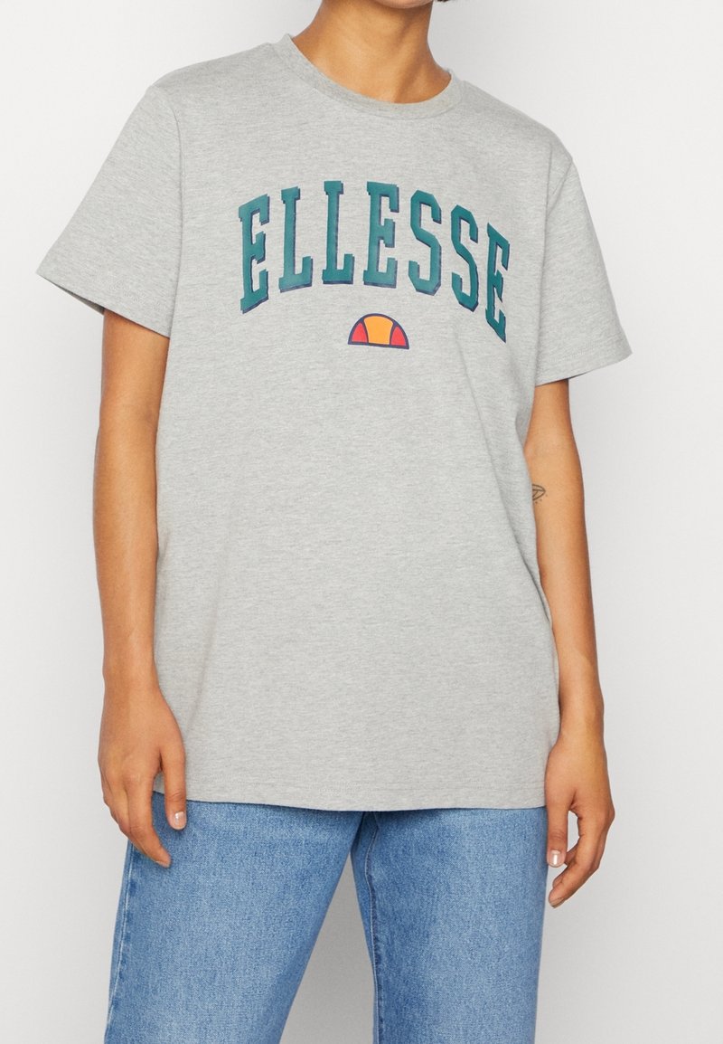 T-shirt en coton gris avec col rond, manches courtes et lettrage audacieux "ELLESSE" en teal et orange, avec un accent en demi-cercle en dessous.