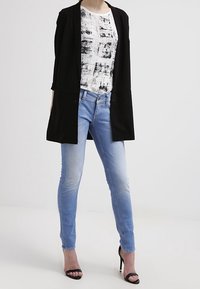 Blazer negro, camiseta gráfica blanca, jeans ajustados de mezclilla azul claro y sandalias de tacón negro. Tejido suave, corte recto, diseño casual.