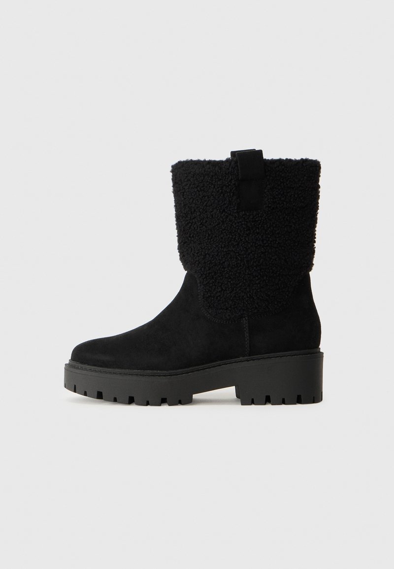 Botas de tornozelo em camurça preta com um superior texturizado semelhante a lã. Apresentam uma sola de borracha grossa com sulcos profundos e uma alça na parte de trás para facilitar o calçar.