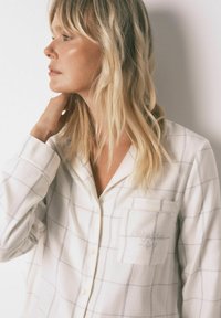 Hvidt ternet pyjamasoverdel med en lomme, der har delikat syning. Materialet har en glat tekstur, med knapper nedad foran.