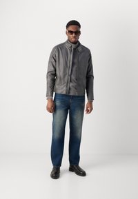 Grauer Lederjacke mit hohem Kragen, Reißverschluss und gerippten Akzenten auf den Schultern, kombiniert mit einem dunklen Hemd und blauen Jeans. Schwarze Stiefel.