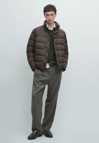 Giacca a vento marrone con colletto alto, indossata sopra un maglione verde scuro e una camicia bianca, abbinata a pantaloni grigi e scarpe nere.