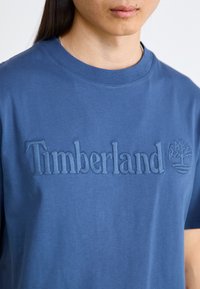 Μπλε βαμβακερό μπλουζάκι με ανάγλυφο λογότυπο "Timberland" και έμβλημα δέντρου σε αντίστοιχο μπλε χρώμα, με στρογγυλή λαιμόκοψη και κοντά μανίκια.