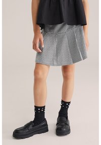 Fekete rövid ujjú felső houndstooth mintás szoknya felett. Fekete platform loaferek fehér gyöngyszerű díszítésű fekete zoknikkal viselve.