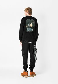 Sudadera negra con estampado gráfico en la parte posterior, combinada con pantalones joggers negros con bolsillos cargo. El modelo lleva botas marrones con cordones.