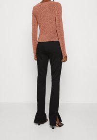 Kvinna som står, klädd i en figursydd brun långärmad topp, svarta högmidjade byxor med bakfickor och svarta klacksandaler.