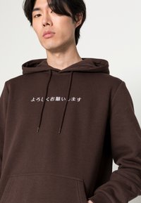 Brun hættebluse med hvid tekstdesign på forsiden. Har en kengurutaske og snore til justerbar pasform. Blød tekstur.