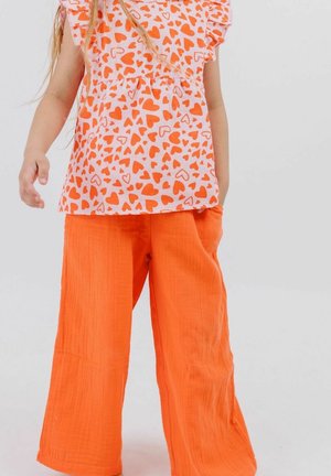Child wearing orange wide-leg pants and a white top with an orange heart pattern, standing against a plain light background.