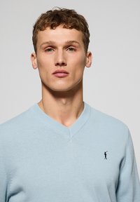 Jasnoniebieski sweter w serek wykonany z miękkiego materiału z prążkowanym dekoltem. Ma małe czarne haftowane logo po lewej stronie klatki piersiowej.