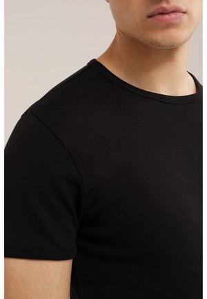 Nahaufnahme einer Person, die ein einfarbiges schwarzes T-Shirt mit kurzen Ärmeln trägt, wobei die obere Brust, die Schulter und ein Teil des Gesichts sichtbar sind.