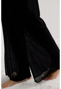 Blote voeten die op zacht wit tapijt stappen, gekleed in vloeiende zwarte broek van geribbelde stof met semi-transparante panelen.