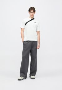 Λευκό βαμβακερό t-shirt με κοντά μανίκια και λογότυπο στο στήθος, σε συνδυασμό με σκούρα γκρι παντελόνι jogger με φαρδιά πόδια και γκρι αθλητικά παπούτσια με μαύρες λεπτομέρειες.