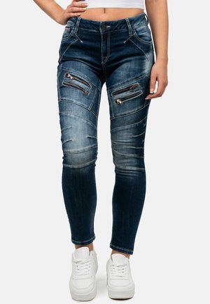 Skinny jeans in dark blue denim with distressed details, diagonal stitching, and four zippered pockets. Paired with white sneakers.