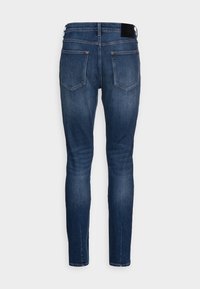 Mörkblå slim-fit jeans visade från baksidan med två bakfickor och en svart varumärkeslapp på midjebandet.