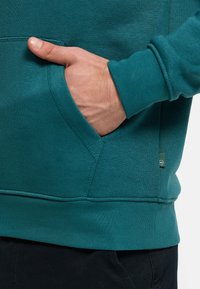 Sudadera con capucha azul verdoso con bolsillo canguro, puños y dobladillo acanalados. Textura suave, con una pequeña etiqueta con el logo en el lateral para la marca.