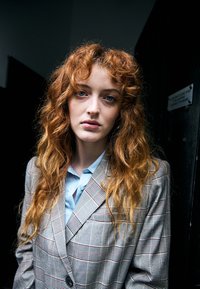 Jeune femme aux longs cheveux roux bouclés et aux yeux bleus porte un blazer à carreaux sur une chemise bleu clair sur un fond sombre.
