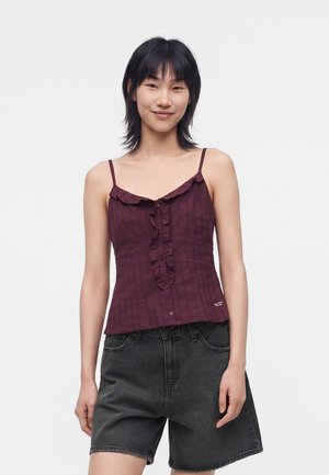 Junge Frau, die ein ärmelloses dunkelviolettes Rüschenoberteil und lockere schwarze Denim-Shorts trägt, steht vor einem schlichten weißen Hintergrund.