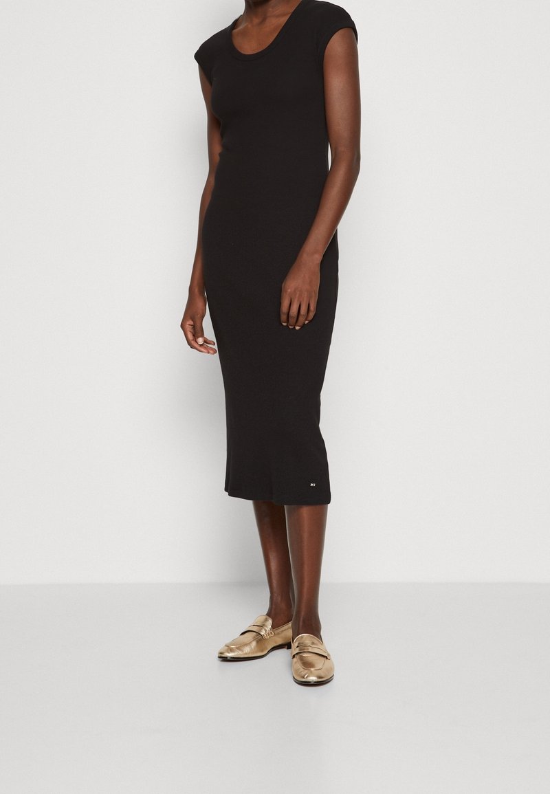 Femme portant une robe midi noire sans manches et des mocassins dorés métalliques, debout devant un fond blanc uni.