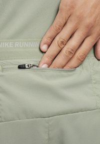 Os calções de corrida da Nike apresentam um tecido verde claro com um padrão de grelha subtil, um bolso com fecho e o nome da marca impresso acima em branco.