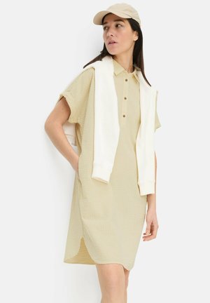 Kvinde iført en beige, kortærmet, stribet kjole, en cremefarvet kasket og en hvid sweatshirt lagt over skuldrene, stående foran en hvid baggrund.