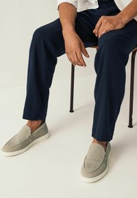 Világosszürke slip-on cipők textúrázott felsőrésszel és fehér talppal, tengerészkék nadrággal párosítva, egy széken pihenve.