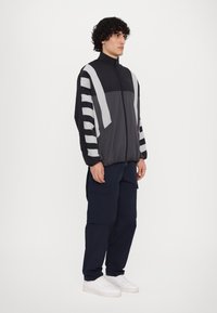 Sort og grå windbreaker med høj krave, med store stribede ærmer og lynlås foran, parret med marineblå cargo-bukser og hvide sneakers.
