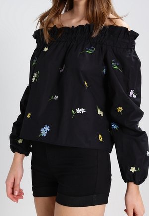 Vrouw draagt een zwarte off-shoulder blouse met kleurrijk geborduurde bloemen, gecombineerd met zwarte shorts met omgeslagen zoom.