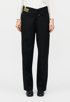 Μπροστινή όψη ενός ατόμου που φοράει φαρδιά μαύρη παντελόνα με ανοιχτή μέση, συνδυασμένη με μαύρα παπούτσια και λευκό μακρυμάνικο μπλουζάκι.