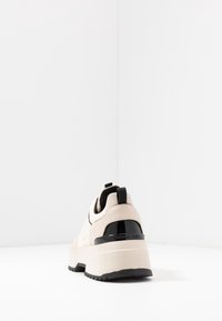 Sneaker beige con tomaia liscia, accenti neri a contrasto, suola spessa bianca e dettaglio del tallone nero lucido. Forma arrotondata con linguetta.