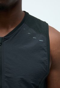 Schwarzes ärmelloses Sporttop mit strukturierten Schulterpaneelen, dezenten reflektierenden Akzenten und glatter Stoffoberfläche. Reißverschluss vorne.