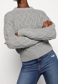 Osoba o ciemnej karnacji nosząca jasnoszary sweter z splotem warkoczowym, z ściągaczami na rękawach i u dołu, w połączeniu z niebieskimi dżinsami w średnim odcieniu.