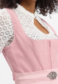 Robe en velours côtelé rose avec un corsage ajusté, de larges bretelles et une fermeture éclair frontale. Soulignée d'une broche argentée décorative sur la ceinture.