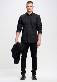 Camisa negra de botones, silueta ajustada, material de algodón, combinada con jeans de mezclilla negros. Los accesorios incluyen una chaqueta negra sostenida en la mano.