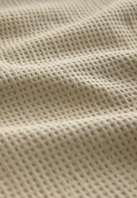 Beiger, strukturierter Stoff mit einem Rastermuster, das erhabene Quadrate zeigt und ein luftiges, weiches Erscheinungsbild mit sichtbaren Wellen erzeugt.