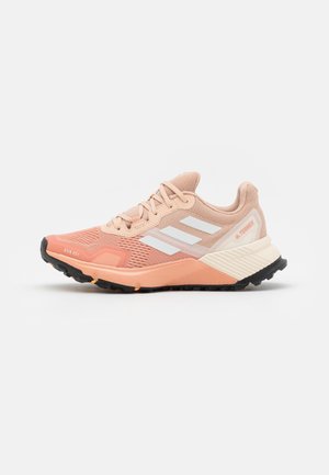 Zapatilla de trail running Adidas Terrex en coral claro y beige, con rayas blancas, suela exterior negra y parte superior de malla para mayor transpirabilidad.