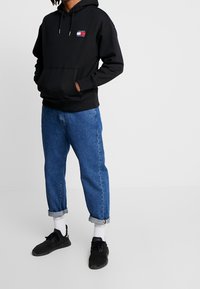 Sudadera negra con capucha y bolsillo frontal, logo en rojo y blanco. Combinada con vaqueros azules con dobladillo y zapatillas negras. Calcetines blancos visibles.