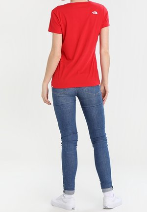 Femme portant un t-shirt rouge avec un petit logo blanc sur le haut du dos, un jean skinny bleu, des chaussettes blanches et des baskets blanches, debout de dos.