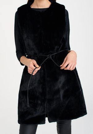 Kobieta nosząca czarną kamizelkę z futrzanym materiałem bez rękawów, z cienkim czarnym paskiem związanym w talii, na czarnej bluzce z długim rękawem i czarnych spodniach.