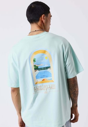 Hombre con camiseta azul claro con un gráfico en la espalda que muestra una vista junto a la piscina a través de una ventana arqueada, palmeras, yate y sol.