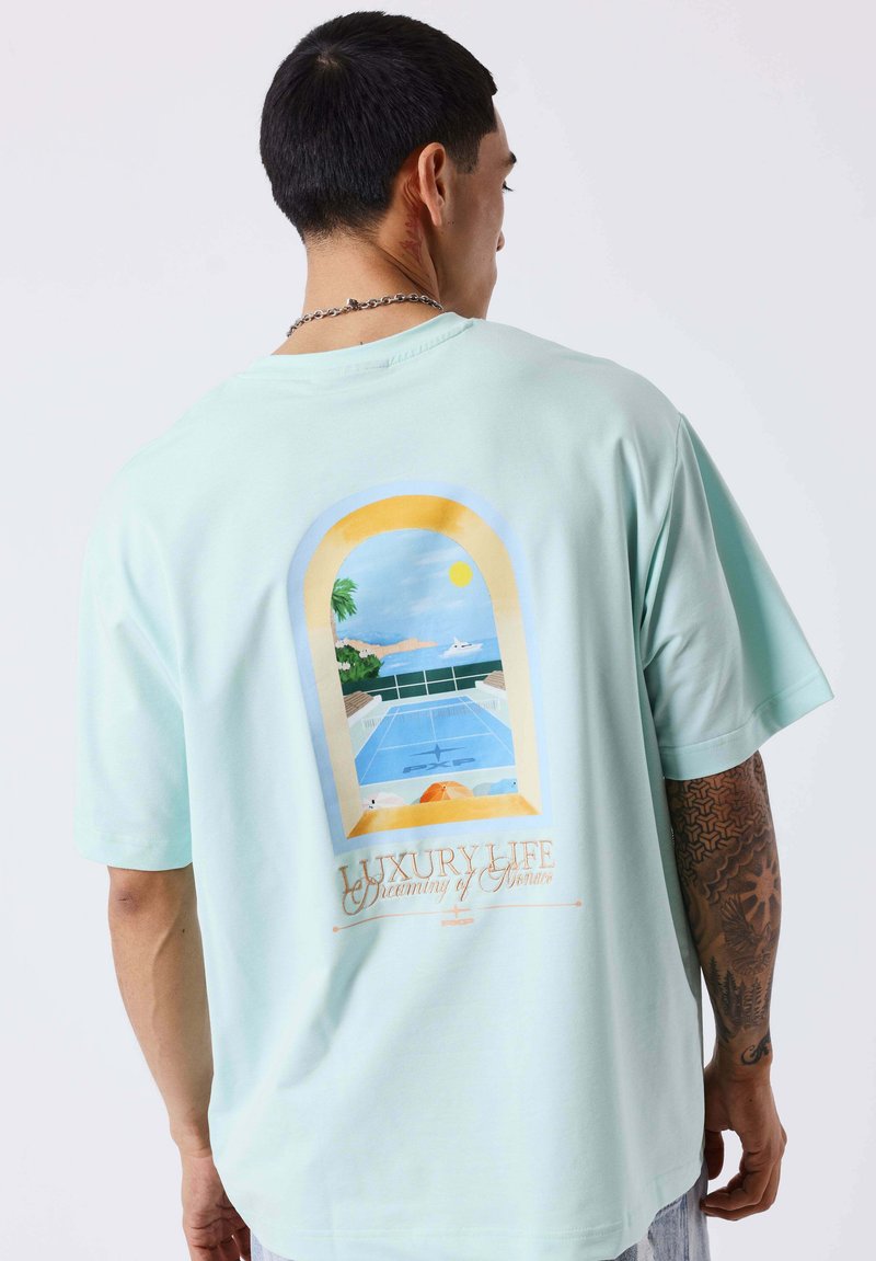 Homme portant un T-shirt bleu clair avec une illustration au dos d'une vue au bord de la piscine à travers une fenêtre en arc, des palmiers, un yacht et le soleil.
