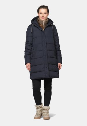Woman wearing long dark blue padded coat with hood, beige fleece high-top boots, and black leggings, standing against plain background.