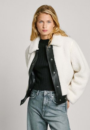 Witte donzige stoffen jas met zwarte leren accenten, drukknoopsluiting en kraaghalslijn, gedragen over een geribbelde zwarte sweater en lichte spijkerbroek.