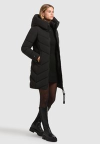 Zwarte winterjas met een hoge kraag, chevronpatroon en ritssluiting. Voorzien van zijzakken en een logo-accent op de ritssluiting.