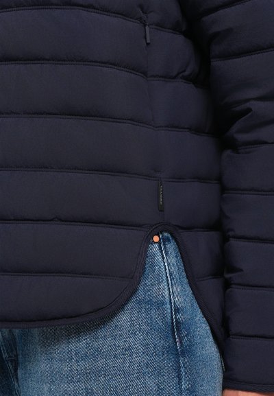Dark blue quilted jacket zipped up, worn over light blue denim jeans, focusing on the lower torso and right arm area.
