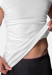 White cotton tank top being lifted to reveal the waistband of black underwear with a three-stripe accent in red, white, and blue.