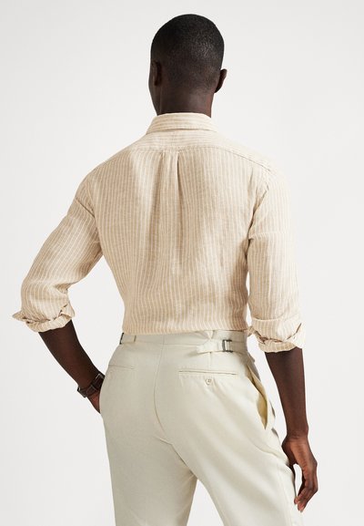 Άνδρας που φοράει μπεζ ριγέ πουκάμισο με μακριά μανίκια γυρισμένα και παντελόνι εκρού, στέκεται πλάτη στην κάμερα.